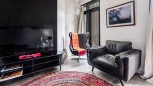 A modern living room with a flat-screen TV, a black leather sofa, a black armchair with an orange throw, a colorful woven rug, and a framed picture on the wall. - Noir Haven Stylish CBD Escape with Private Rooftop (Melbourne)