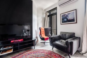 A modern living room with a flat-screen TV, a black leather sofa, a black armchair with an orange throw, a colorful woven rug, and a framed picture on the wall. - Noir Haven Stylish CBD Escape with Private Rooftop (Melbourne)