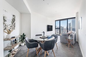 Ein modernes Wohnzimmer mit einem runden Glastisch, grauen Polsterstühlen, einem Flachbildfernseher und Blick auf die Stadt durch große Fenster.