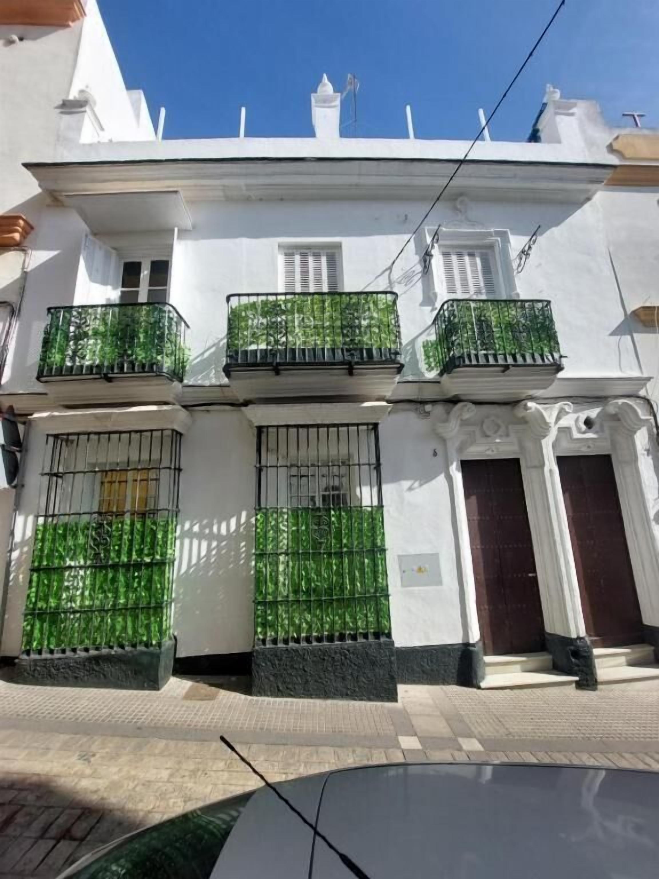 A white building with three balconies, each with metal railings and a green plant. There are two doors and a window on the facade.