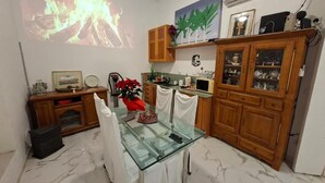 A dining area with a glass table, chairs, a projector displaying a fire, wooden cabinets, and a wall-mounted shelf with decorative items.
