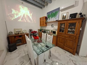 Un comedor con mesa de vidrio, sillas, un proyector que muestra fuego, armarios de madera y un estante de pared con objetos decorativos.
