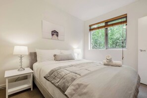 A neatly arranged bedroom with a bed, bedside lamps, a window with a view of greenery, and a framed picture on the wall. - Experience the St Kilda Vibe from a Cosy Retreat (Saint Kilda)