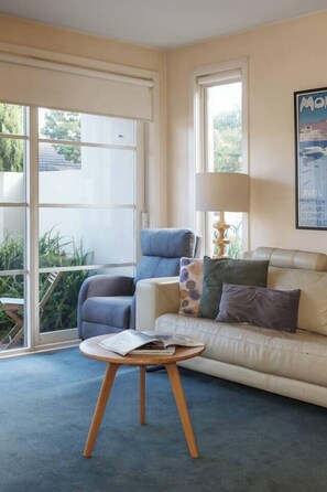 Un salon comprenant un canapé, un fauteuil, une petite table ronde avec des magazines, une lampe et une fenêtre donnant sur un paysage verdoyant.