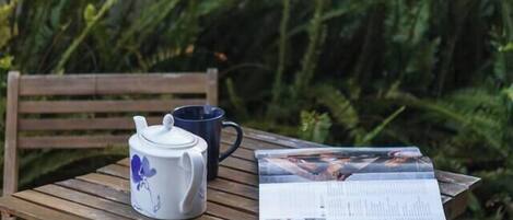 A wooden table with a white teapot and a blue cup, a newspaper, and a chair.