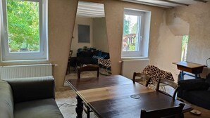 A room with a dining table, chairs, a sofa, and two windows.