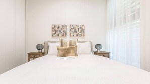Uma cama bem arrumada com edredom branco e almofadas listradas, ladeada por duas mesas de cabeceira com abajures, em um quarto com parede branca lisa e uma grande janela com cortinas finas.