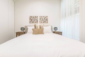 A neatly made bed with a white comforter and striped pillows, flanked by two bedside tables with lamps, in a room with a plain white wall and a large window with sheer curtains. - Alexandra Elegance Riverside Living in Style (South Yarra)