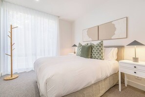 A hotel room with a large bed, a bedside table with a lamp, a coat rack, and a window with sheer curtains. - Alexandra Elegance Riverside Living in Style (South Yarra)