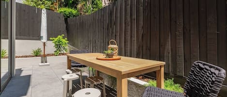 An outdoor patio with a wooden table and four chairs, surrounded by a wooden fence and greenery.