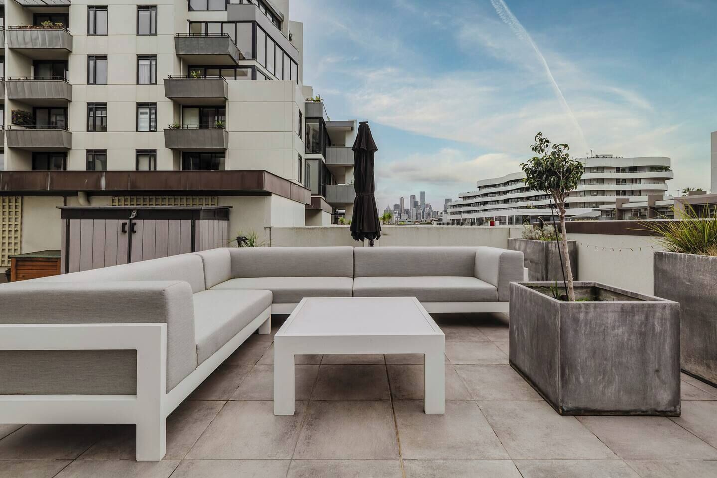 Une terrasse sur le toit aménagée avec un canapé d’angle, une table basse blanche et un bac à plantes en béton dans lequel est planté un arbre.