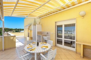 Une terrasse extérieure couverte, équipée d’une table pour les repas, d’un évier intégré et d’une porte-fenêtre coulissante donnant sur un espace intérieur.