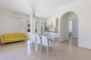 Eine moderne Küche mit weißen Schränken, ein Essbereich mit weißem Tisch und Stühlen und ein gelbes Sofa in einem hellen Wohnraum.