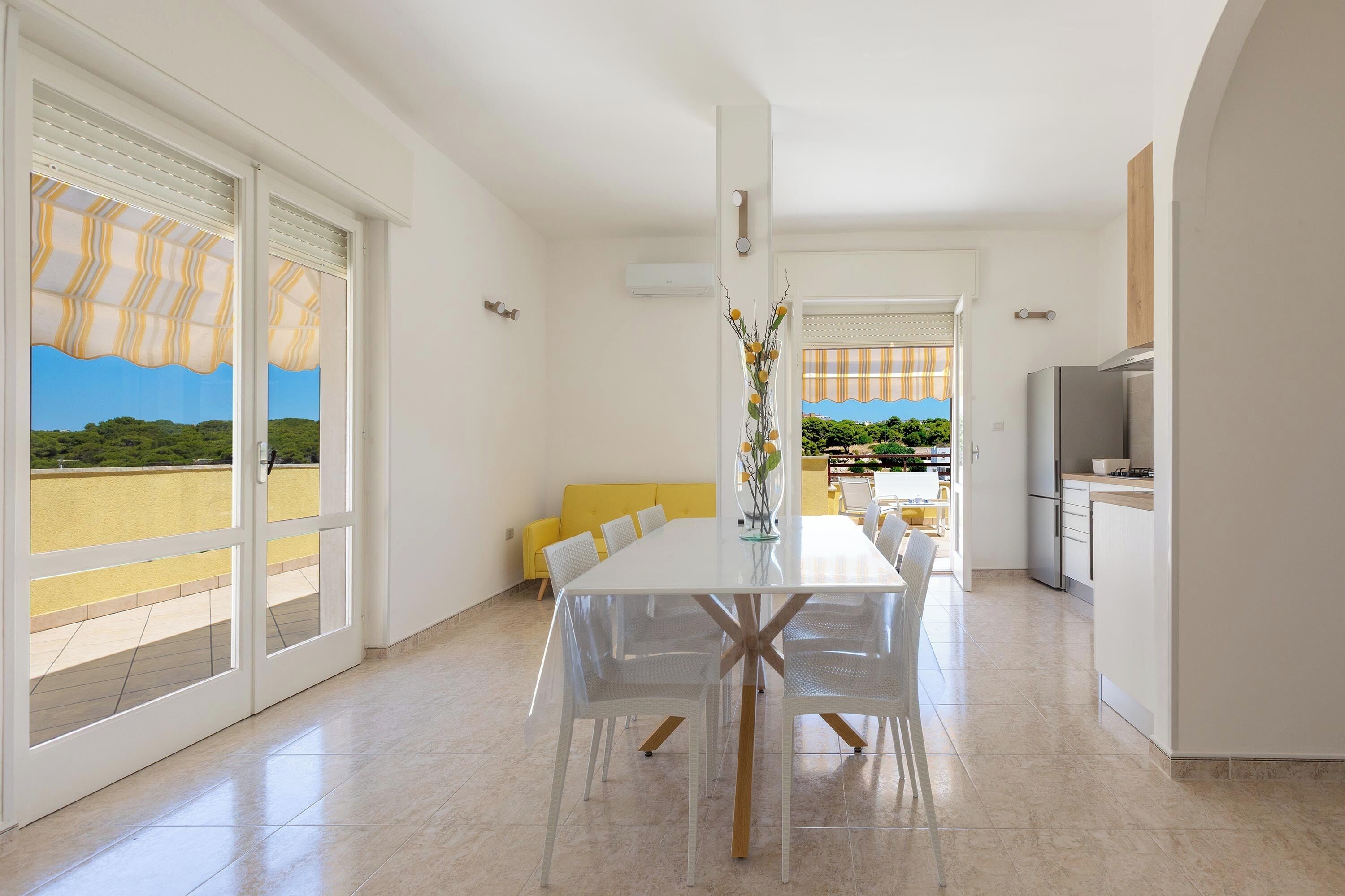 A modern kitchen with a dining area, large windows, and a view of greenery.