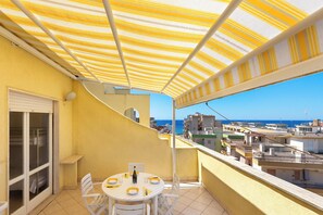 Un balcon agrémenté d’un auvent à rayures jaunes, une table ronde avec des chaises, et une vue sur la mer et les bâtiments.