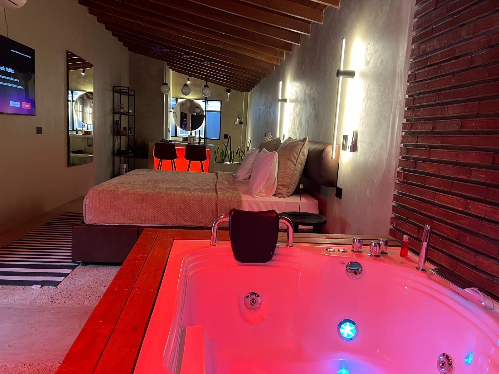 A modern bedroom with a large bed, a sitting area with a red chair, a large bathtub with lights, and a TV on the wall.