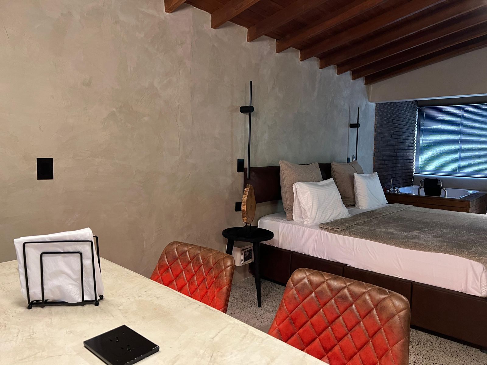 A hotel room with a bed, a desk, and a chair. There is a window with blinds, a wall-mounted lamp, and a small table with a chair.