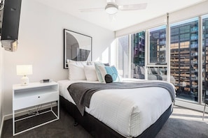 Een moderne slaapkamer met een groot bed, een nachtkastje, een televisie en een raam met uitzicht op de stad.