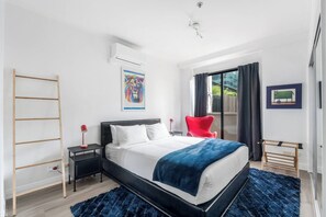 A modern bedroom with a bed, a red chair, a wooden ladder, and a window with curtains. - The New York Edit CBD Living with Private Rooftop (Melbourne)
