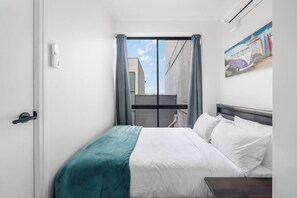 A hotel room with a bed, a nightstand, a window with curtains, and a painting on the wall. - The New York Edit CBD Living with Private Rooftop (Melbourne)