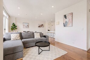 Ein modernes Wohnzimmer mit einem grauen Ecksofa, einem runden Couchtisch und einem kleinen Teppich. Im Hintergrund ist ein Essbereich mit einer Bar und Hockern zu sehen.