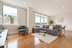 A modern living room with a grey sectional sofa, a small round coffee table, and a grey armchair. There is a dining area with a table and chairs, and a large window offering a view of buildings. - Cityscape Retreat Modern Comfort near Yarra River (Southbank)