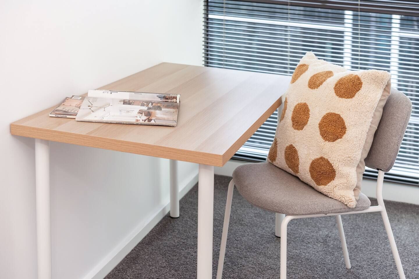 A small, modern room with a wooden table, a grey chair with a polka-dotted cushion, and a window with blinds.