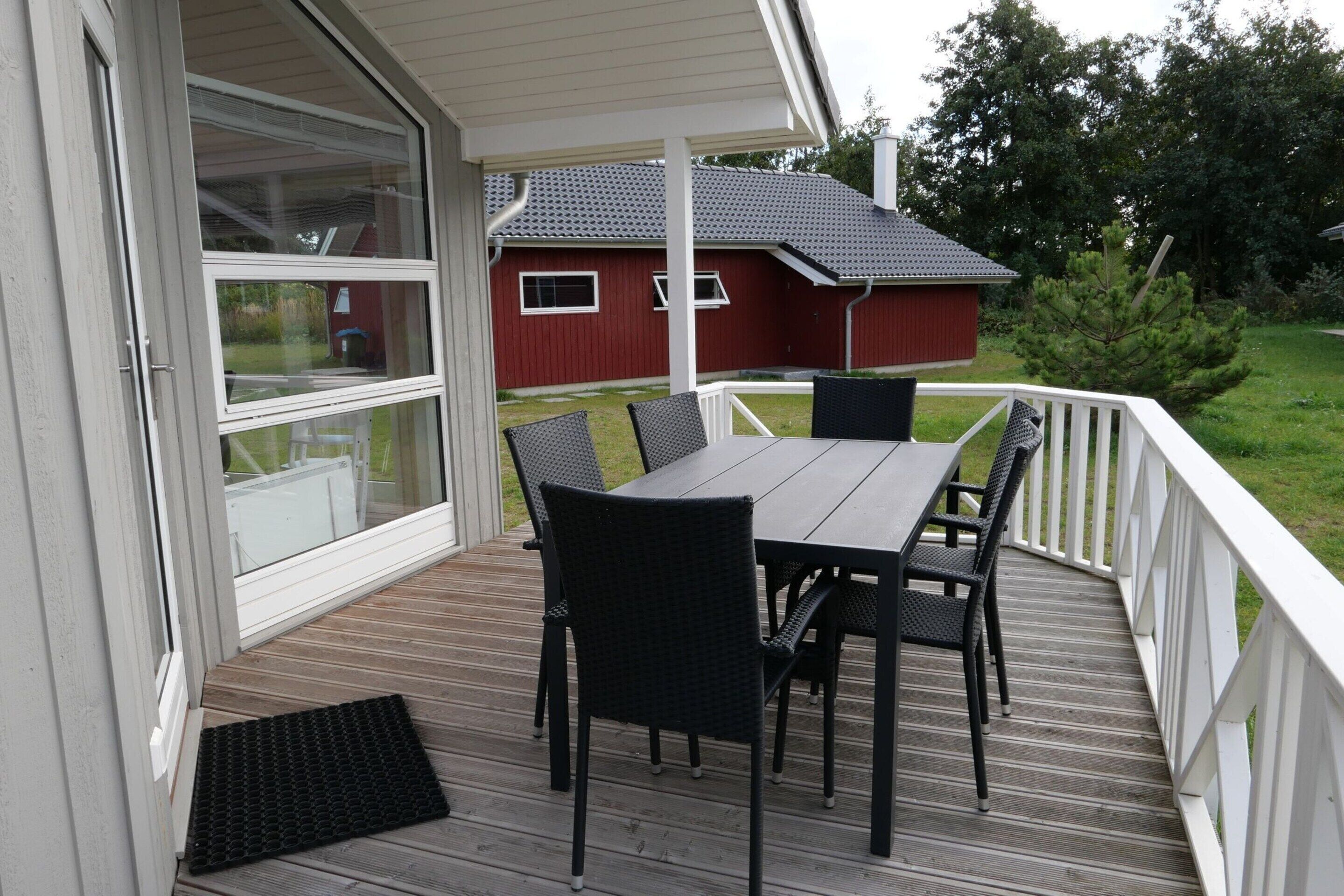 Eine überdachte Terrasse mit Tisch und Stühlen, ein rotes Bauernhaus im Hintergrund und eine weiße Geländer.