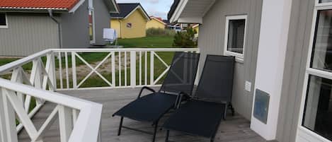 A covered outdoor area with a wooden deck, two black lounge chairs, a white railing, and a view of neighboring houses.