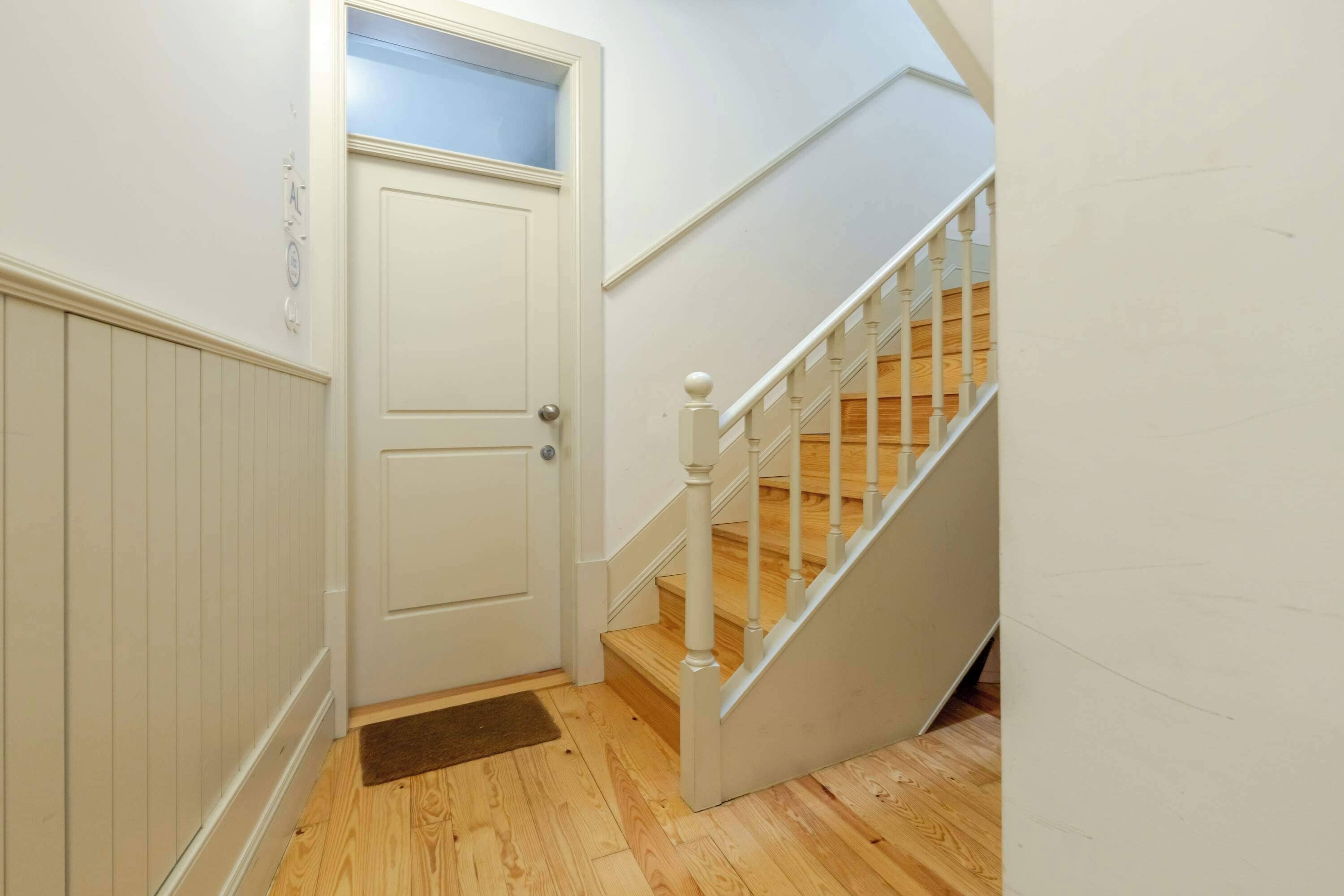 A hallway with a white door, a wooden floor, and a white staircase with a wooden handrail.