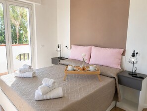 Ein ordentlich gemachtes Bett mit einem Frühstücksbrettchen, ein Nachttisch mit Lampe und Blick auf Grün hinter dem Fenster.