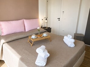 A hotel room with a bed, a bedside table with a lamp, a tray with coffee cups and croissants, and rolled towels on the bed. - Riace Mare – Premium | 100 m from the beach (Riace Marina)