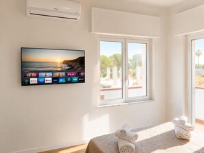 Ein Hotelzimmer mit einem wandmontierten Flachbildfernseher, der verschiedene Streaming-Dienste anzeigt, einem großen Fenster mit Blick auf Palmen und einem Bett mit zusammengerollten Handtüchern.
