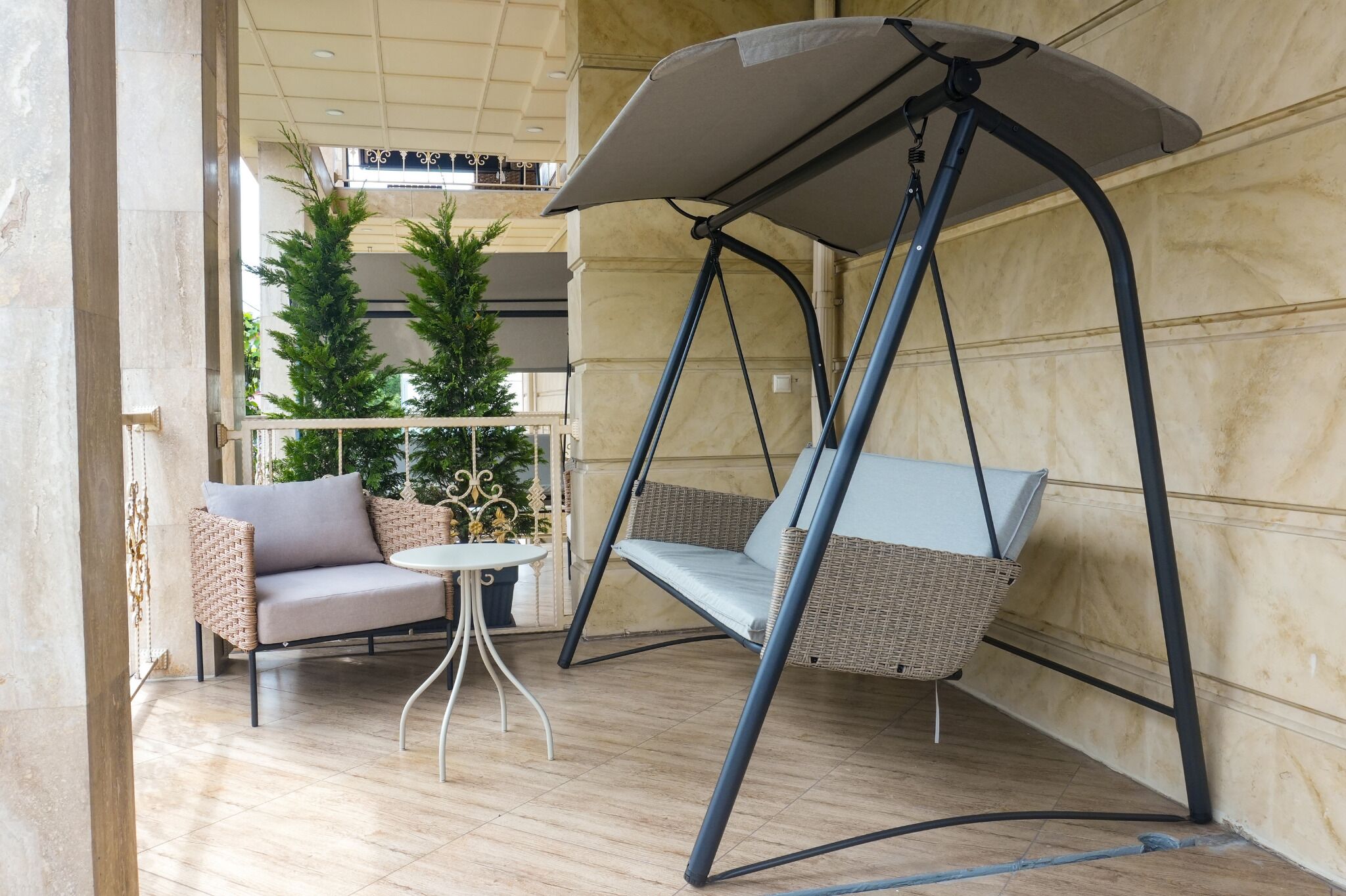 A swing chair with a canopy, a wicker sofa, and a small round table in a room with tiled flooring and a wooden wall.