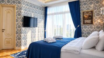 A hotel room with a bed, a flat-screen TV, a large window with blue curtains, a chandelier, and a framed picture on the wall.
