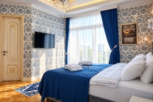 Habitación de hotel con cama, televisor de pantalla plana, ventana grande con cortinas azules, candelabro y un cuadro en la pared.