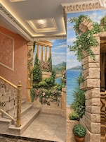 Una imponente escalera con barandillas ornamentadas y un mural que representa una escena clásica con columnas y un paisaje.