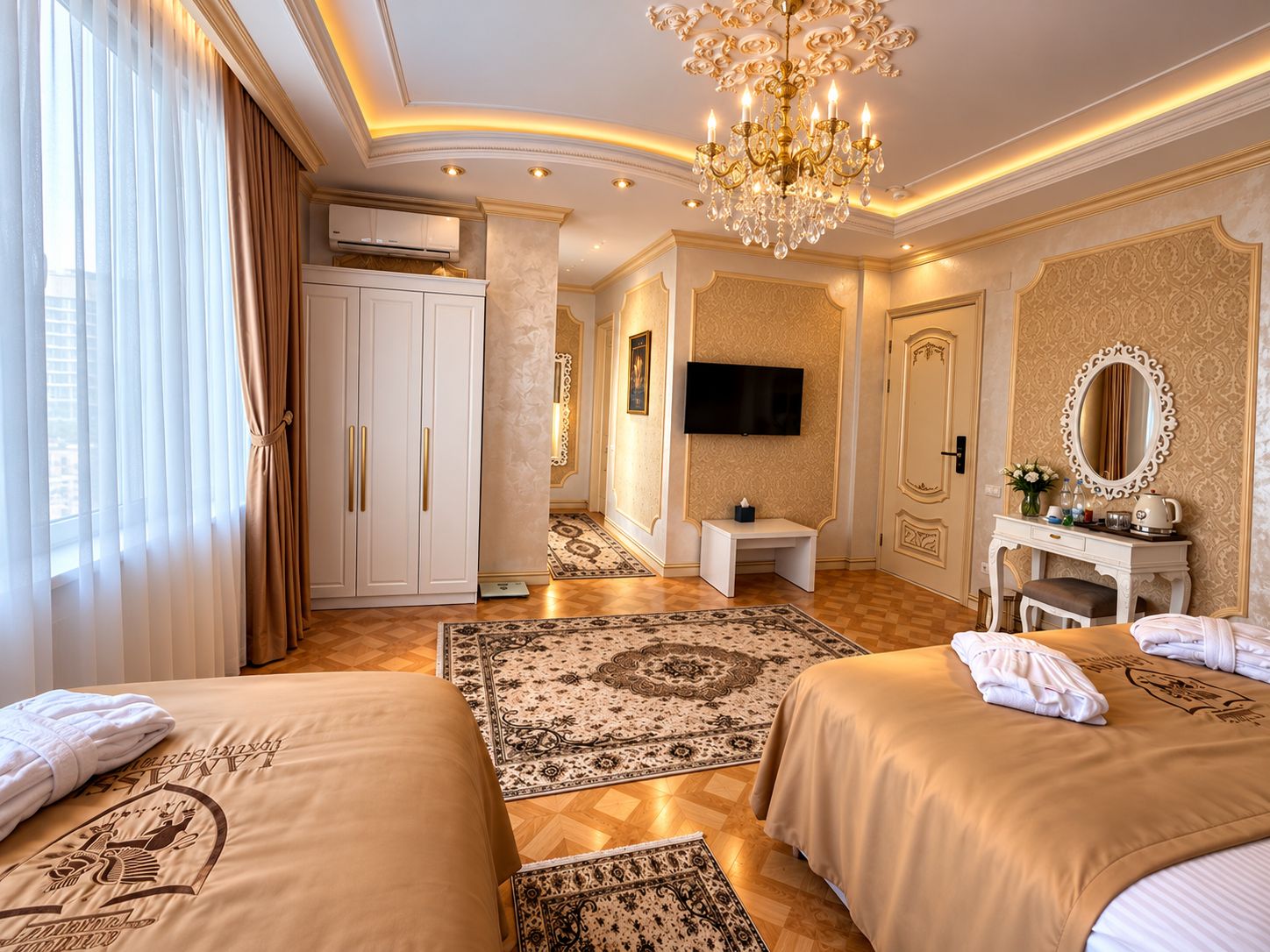 A hotel room with two beds, a television, a desk, a mirror, and a chandelier.