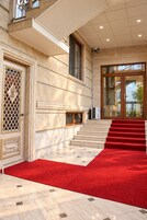 Entrada de hotel con paredes de mármol, alfombra roja y una escalera que lleva a una puerta de cristal.