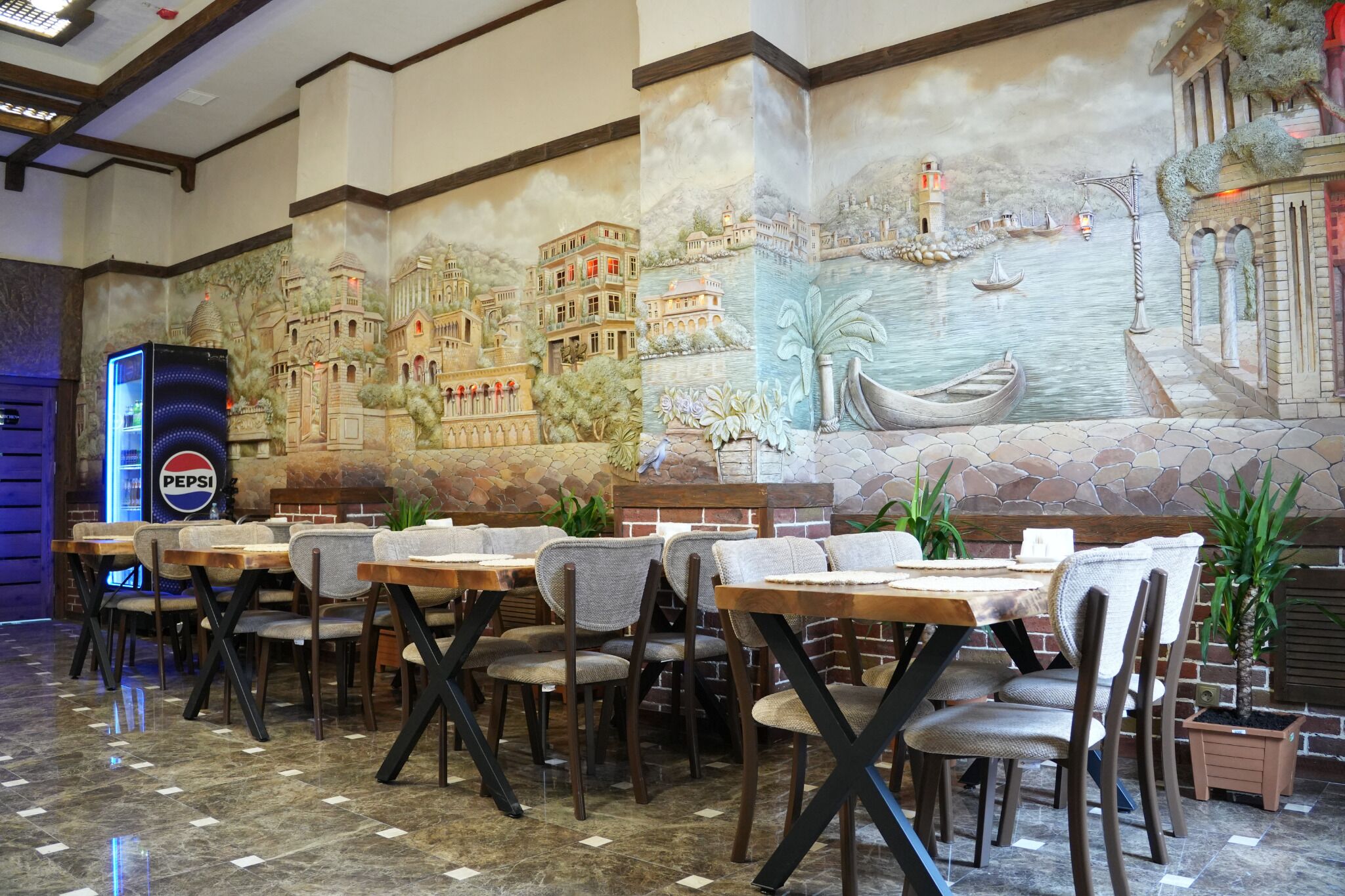 Un restaurante con un mural de una escena costera, mesas con sillas y un refrigerador de Pepsi.