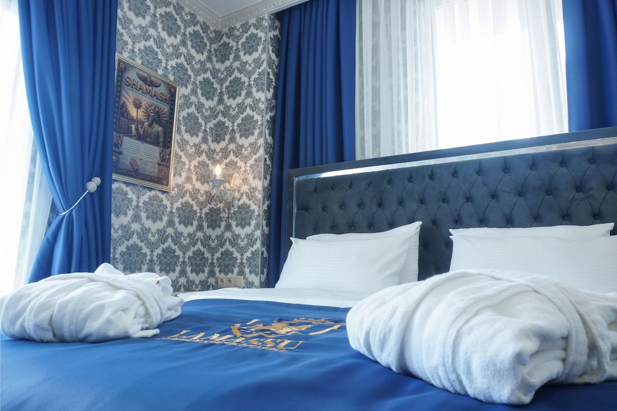 A hotel room with a large bed, blue curtains, a wall-mounted lamp, and a framed picture.