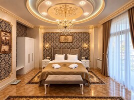 A luxurious bedroom with a large bed, ornate wallpaper, a chandelier, and a view of the outdoors.