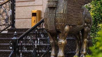 A bronze statue of a winged creature with a detailed headdress, positioned on a pedestal in front of a building with ornate wall decorations and a staircase.