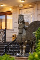 A bronze statue of a winged creature with a detailed headdress, positioned on a pedestal in front of a building with ornate wall decorations and a staircase.
