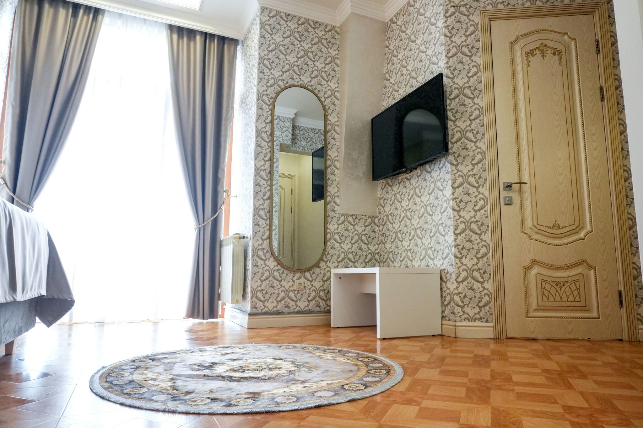 Una habitación con piso de madera, un tapete redondo con diseño, un escritorio blanco, un televisor montado en la pared, una puerta con detalles ornamentales y una ventana con cortinas.