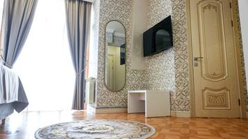 Una habitación con piso de madera, un tapete redondo con diseño, un escritorio blanco, un televisor montado en la pared, una puerta con detalles ornamentales y una ventana con cortinas.