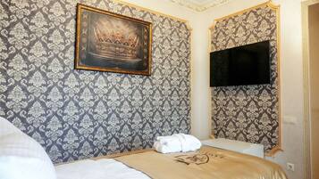 Ein Hotelzimmer mit ornamentalem Wandpapier, einem gerahmten Kunstwerk einer Königskrone, einem ordentlich bezogenen Bett und einem an der Wand befestigten Fernseher.