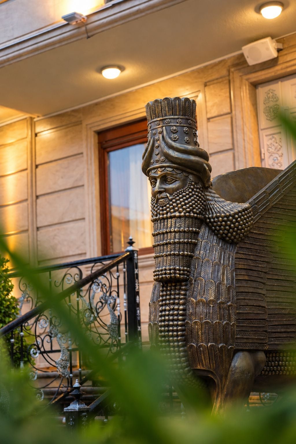 Una estatua de bronce de una figura alada con un tocado detallado, ubicada frente a un edificio con puerta de madera y barandilla metálica.