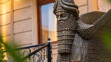 Una estatua de bronce de una figura alada con un tocado detallado, ubicada frente a un edificio con puerta de madera y barandilla metálica.