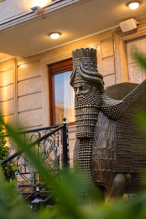 Una estatua de bronce de una figura alada con un tocado detallado, ubicada frente a un edificio con puerta de madera y barandilla metálica.